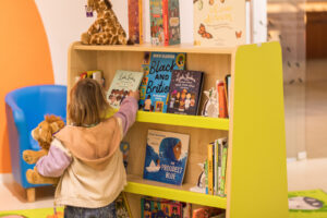 A young girl with dark blonde hair is holding a cuddly toy lion. She is reaching for a book on the yellow bookcase.